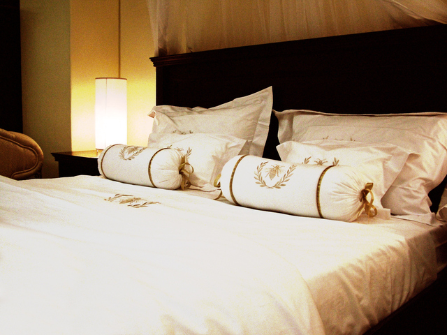 Čierna posteľ s bielym matracom a obliečkami, nočný stolík so zapálenou stolnou lampou.jpg